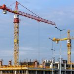 Multiple tower cranes working on a large construction site with blue sky backdrop.