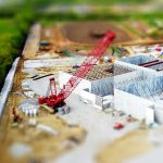 Aerial view of a construction site with a red crane amidst green fields and concrete buildings.